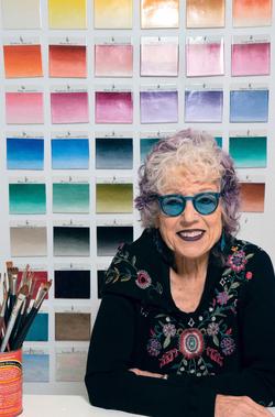 A photo of Judy Chicago sitting in front of a wall of colored swatches.