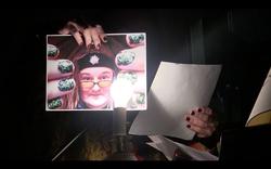 A photo of hands behind a microphone showing a photo of poet CAConrad framed between painted fingernails.