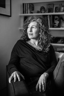 A photo of poet Susan Briante, looking toward a window, with shelving and a framed artwork behind her.