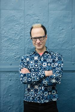 A photograph of poet Wayne Koestenbaum, wearing a blue printed shirt, with arms crossed in front of a blue wall.