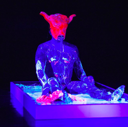 Ron Athey, Acephalous Monster, 2019, MoCA Skopje. Photo: Andreja Kargačin. [The artist Ron Athey, wearing a minotaur mask, is seated in a shallow box of viscous fluids under a blacklight in shades of blue, white, and pink, covered in drippy fluids.]