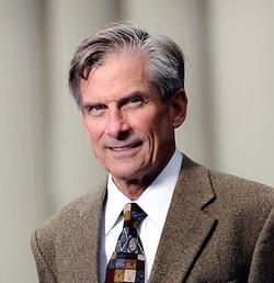 This is a headshot of Artist William D. Adams against an off-white curtain background. Adams wears a dark beige blazer, white shirt, and patterned tie.