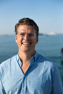 A photo of Ben Jones standing in front of a body of water in a blue shirt, with the warm sunlight shining on his face.