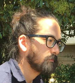 A photograph of Brandon Shimoda, wearing glasses, in front of green leaves and branches.