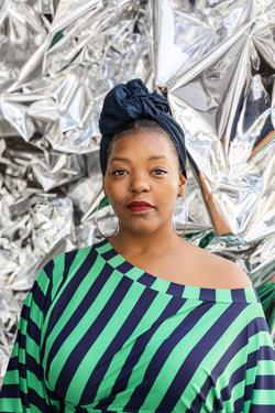 This is a photo of Poet and Activist, Mahogany L. Browne against a metallic background. She's wearing a green and dark blue striped shirt, off the shoulder, a navy blue head wrap, and large gold hopped earrings. 
