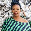 This is a photo of Poet and Activist, Mahogany L. Browne against a metallic background. She's wearing a green and dark blue striped shirt, off the shoulder, a navy blue head wrap, and large gold hopped earrings. 