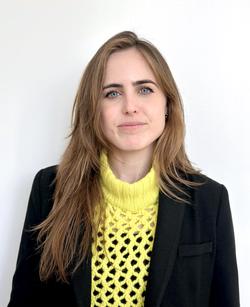 A portrait of writer Cassie Packard in front of a white background wearing a yellow sweater and black blazer with long blonde hair. 