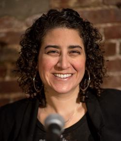 A photograph of Caroline Rothstein, smiling behind a microphone.