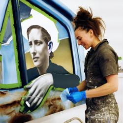 Artist Chloe Chiasson wearing an apron splattered with white paint as she works on an artwork of a person wearing a cowboy hat looking out from the window of a car. The painting is green and blue and the figure is portrayed in cinematic black and white.