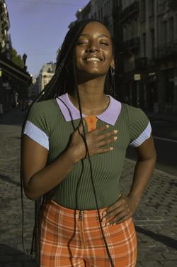 A photo of Yaissa Jiménez smiling with hand on chest, green shirt and orange pants on a city street