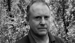 A black-and-white photographic portrait of curator Enrique Juncosa. It is a close up of Enrique's face and shoulders, and he is standing in front of a wooded forest.