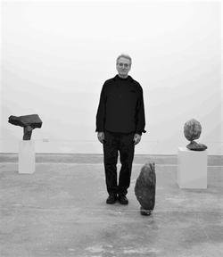 A black and white photo of [George Quasha] among sculptures. 