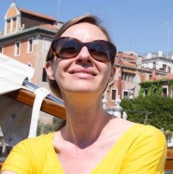 This is a sunny portrait of the Rail's Art Editor, Amanda Gluibizzi with houses in the background and a blue sky. Gluibizzi is wearing a yellow shirt and sunglasses.