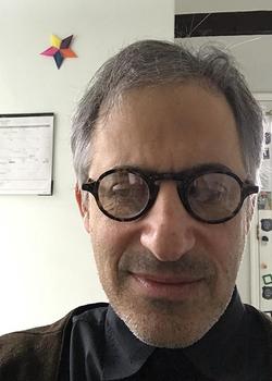 This is a selfie of Artist Max Goldfarb with a background and pictures hanging on the wall. He's wearing black framed glasses.