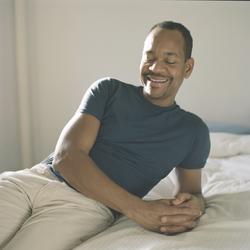 This is a photo of Artist Lyle Ashton Harris in a room leaning on a bed. The background is off-white and Harris's shirt is dark green.