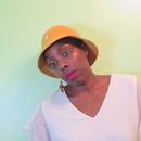 A photo of Joselia Rebekah Hughes, an Afro-Caribbean person in a white T shirt and yellow hat, glasses and red lipstick, body titled slightly, in front of a green hued background. 