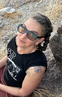 A white Latinx Jewish person with long brown hair in a braid, streaked with silver. They are wearing light-colored shades with blue frames, and have multiple facial piercings. They are sitting on rocky earth, looking up toward the camera, wearing a sleeveless Festival for all Skid Row Artists t-shirt and dusty rose-colored pants. A tattoo of a flowering succulent is visible peeking out of their shirt on their left shoulder. 