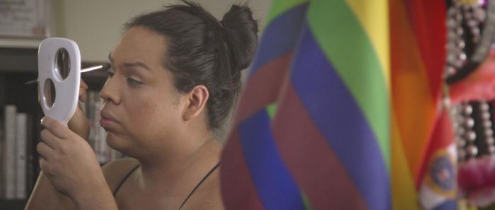 Film still of a person looking in a mirror with a rainbow flag covering half of the righthand side of the image.