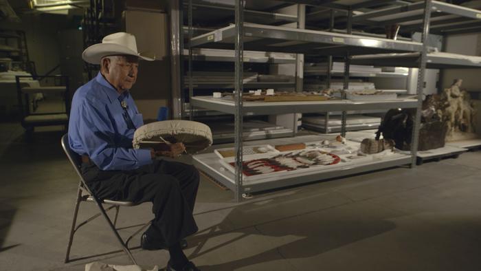 A person sits in a folding chair with a hat on holding a drum in a storage space.