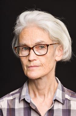 A color photo portrait of the artist Jenny Polak from the shoulders up. Jenny is wearing glasses and has short silver hair. She is wearing a brown and white plaid shirt and staring directly at the camera.