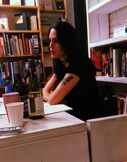 A photograph of poet Kay Gabriel sitting an a desk, near bookshelves, an open notebook, wine bottles and cups.