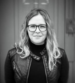 A black and white photograph of curator Laurel V. McLaughlin. She is standing in front of a doorway, wearing a leather jacket and a black turtleneck.