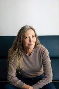 Color-photo portrait of artist Liz Larner, wearing a light brown sweater with crossed arms and seated on a blue bench