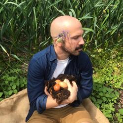 Poet Lucas de Lima with flowers behind their ear, sitting on green ground, holding 3 eggs 