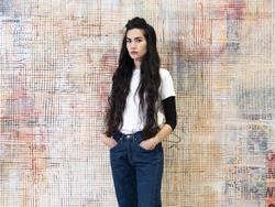 A color photographic portrait of artist Mandy El-Sayegh, standing in front of one of our intricately patterned paintings. Her long brown hair is down, and she is wearing jeans and a white t-shirt.