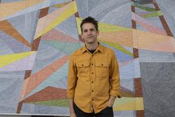 A photograph portrait of artist Matt Phillips from the waist up, standing in front of one of his artworks. He is wearing a yellow shirt and black pants. His hand is in his pocket. 