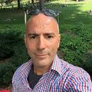 A photo of poet Michael Broder, wearing a button up and glasses on his head, in front of a field of green grass.