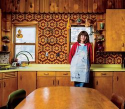 A color photograph of artist Michelle Grabner leaning against a kitchen counter. She's wearing a denim apron in a 1970s kitchen with light wood cabinets and a yellow countertop and a groovy, printed orange wallpaper. She is wearing a red sweater.