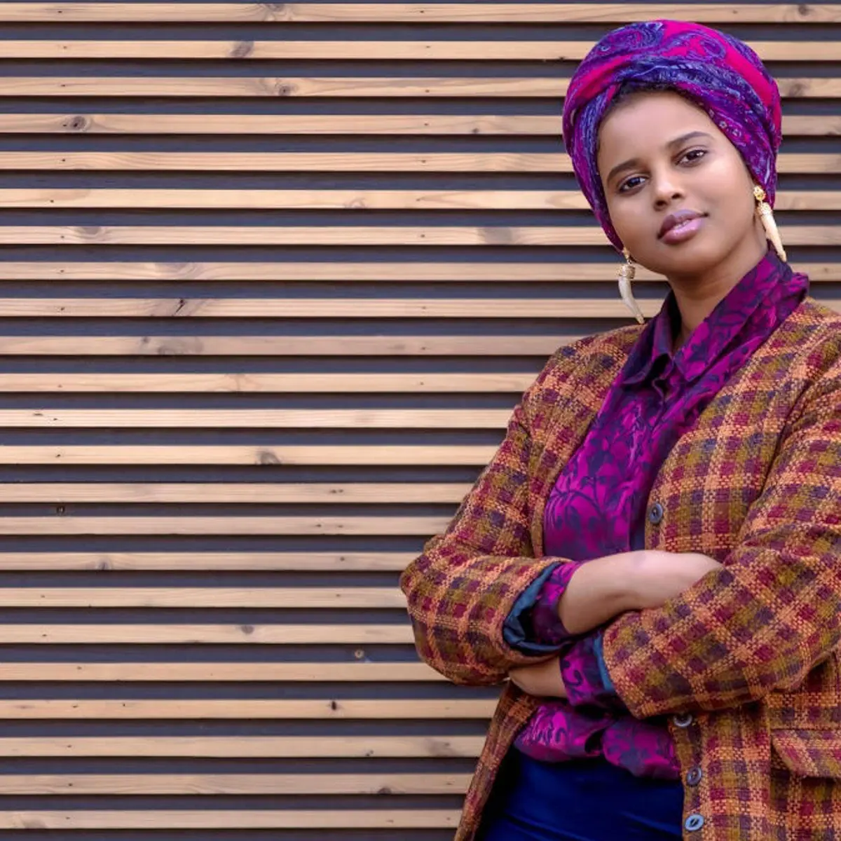 A photo of [Momtaza Mehri] in a patterned blazer and headscarf, arms crossed, against horizontal wood background.  
