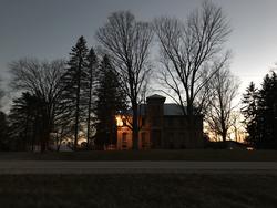 A color photograph of a house in Wisconsin. Its nighttime and the sun is glowing behind the house's rooftop. Its surrounded by trees and a grey, barely sunlit sky.