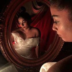 A photograph of poet Ramya Raya Ramana reflected in a round wooden mirror, wearing a white gown.