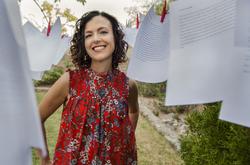 A photo of poet Rosa Alcalá in a red shirt, smiling between sheets of paper hung on a clothesline.