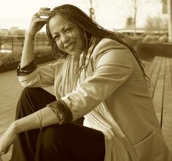 A sepia photograph of poet Samiya Bashir sitting on a long outdoor bench and smiling.