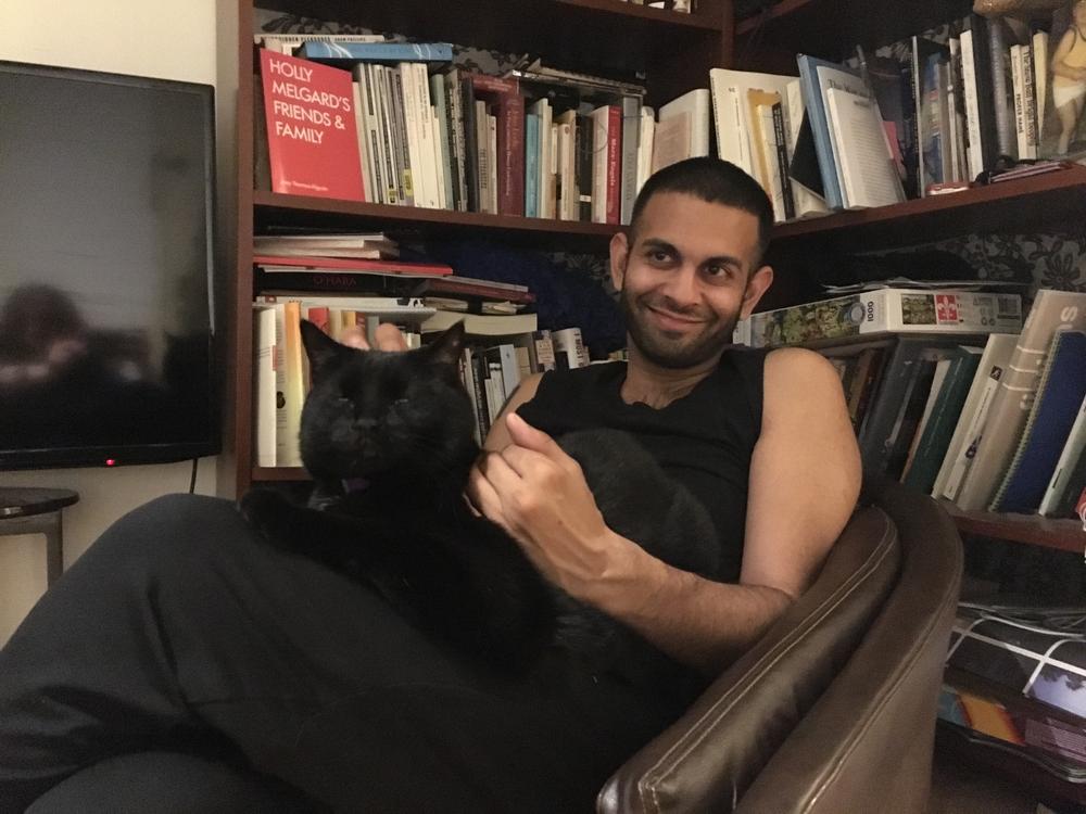 A photo of Shiv Kotecha sitting in front of a bookshelf in a leather chair, with a black cat sleeping on his lap.
