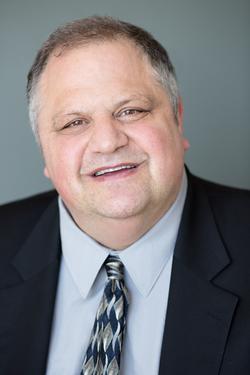 This is a portrait of Writer Steve Silberman with a gray background. He's wearing a dark blazer with a green and blue patterned tie.