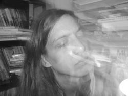 A black and white photo of [Steph Gray] in front of books with their eyes closed, long exposure light over the image.