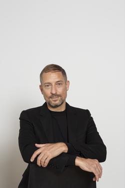 A photographic portrait of the curator Stuart Comer. His arms are crossed and he is wearing a black blazer and shirt. He is standing in front of a plain white background.