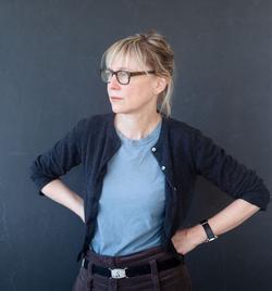 This is a photo of Artist, Suzanne Bocanegra with a dark gray background. She is posing with her hands on hips and angled to her right.