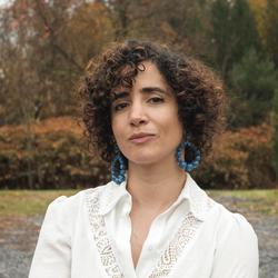 A color photograph of artist Tania El Khoury wearing blue hoop earrings and a white shirt. Tania is standing in an autumnal park, surrounded by grass and trees with orange and green leaves.