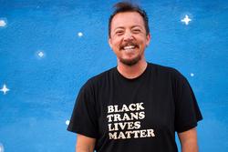 A photo of poet TC Tolbert, wearing a Black Trans Lives Matter shirt in front of a background of painted stars.