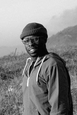 Black and white photography portrait of artist Woody De Othello wearing a sweatshirt and beanie in an open field outdoors. 