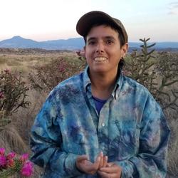 A portrait of Amelia Bande in a colorful blue jacket and baseball cap, smiling, in front of desert brush and mountains in the distance